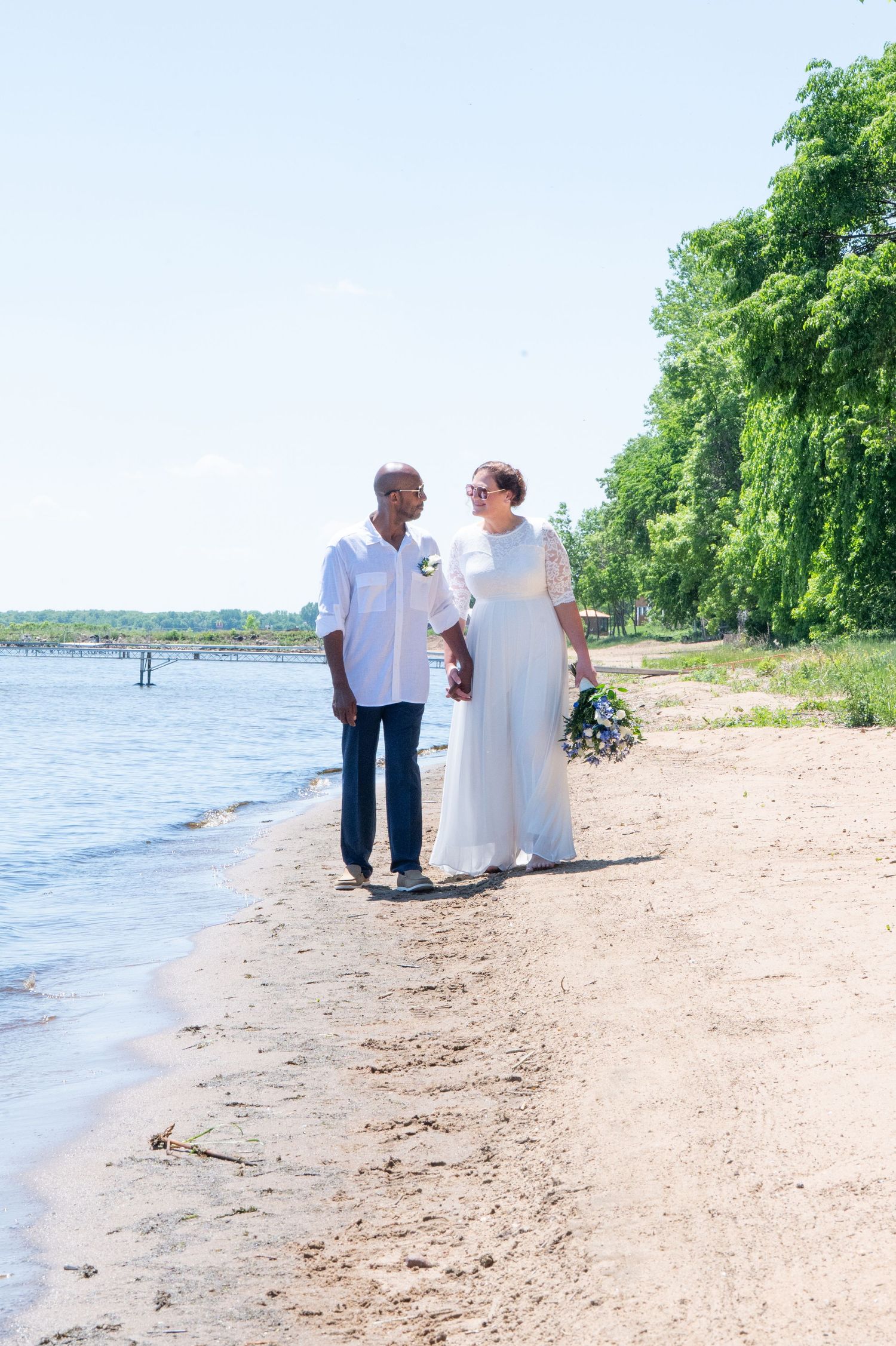 Bride and groom walking hand in hand along a sunny sandy beach by the water.