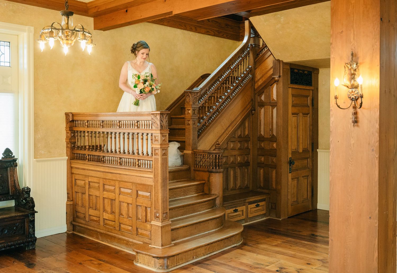 Bride in a white lace gown holding a bouquet stands on an ornate wooden staircase in a historic mansion.