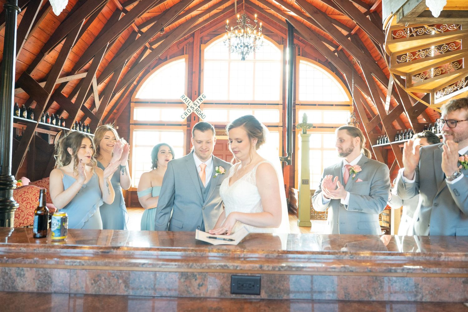 Bride and groom sign marriage documents surrounded by smiling wedding party in a rustic wood-beamed venue.