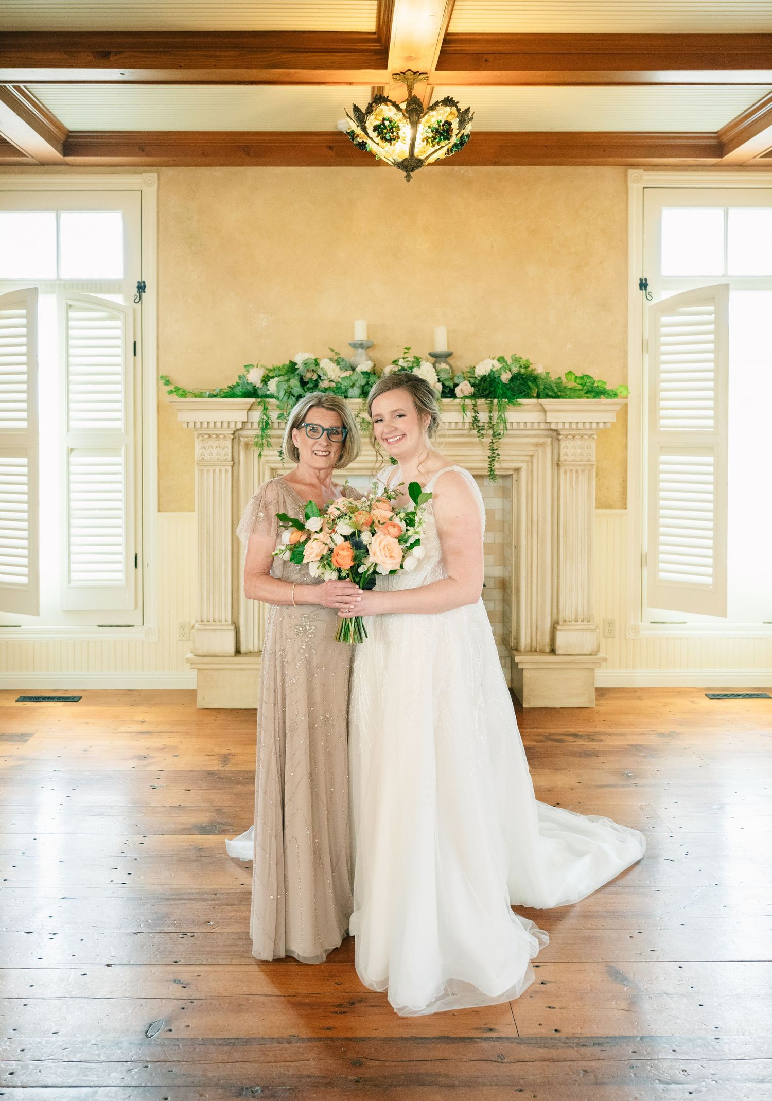 Bride poses with older woman in gold dress before floral fireplace on wood floor.
