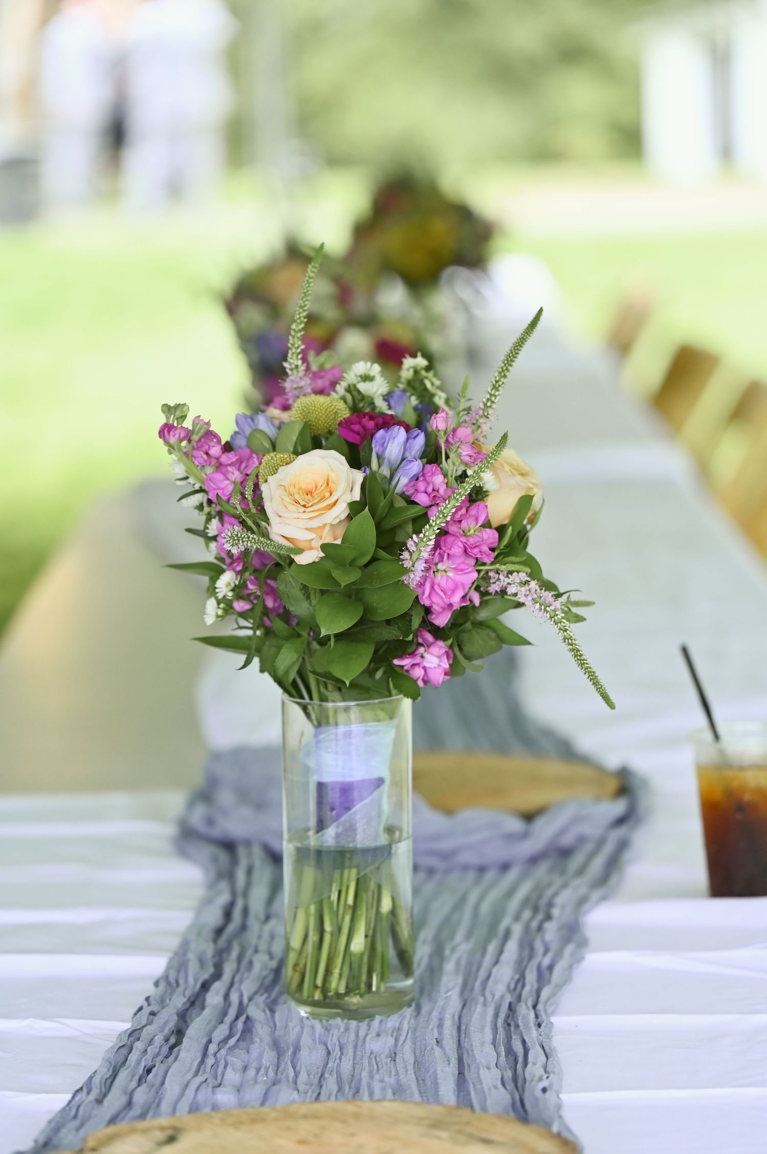 Elegant outdoor wedding table with pink roses and purple wildflowers in a glass vase on a gray linen runner.