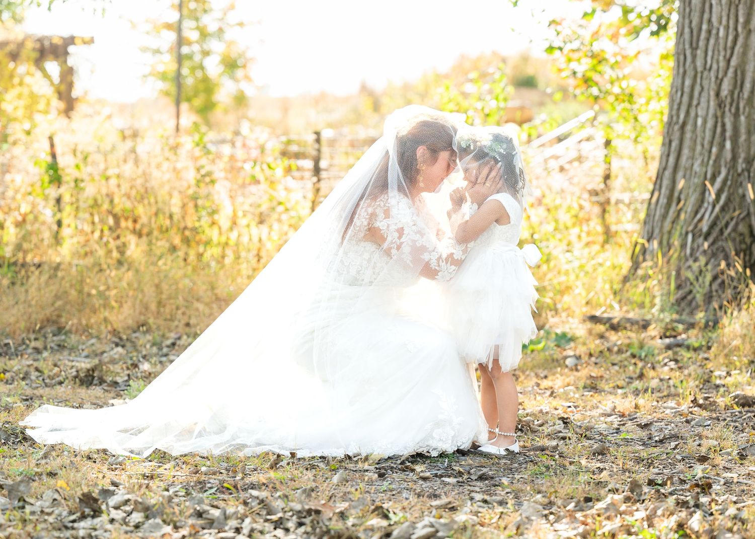 Bride in white gown and veil kneeling outdoors, sharing a tender moment with a young flower girl in autumn setting.