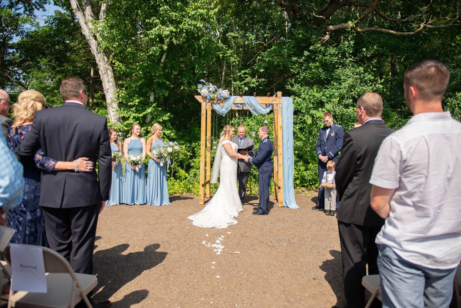 Outdoor forest wedding ceremony with wooden arch and light blue drapery surrounded by guests. Zimmerman MN Weddings