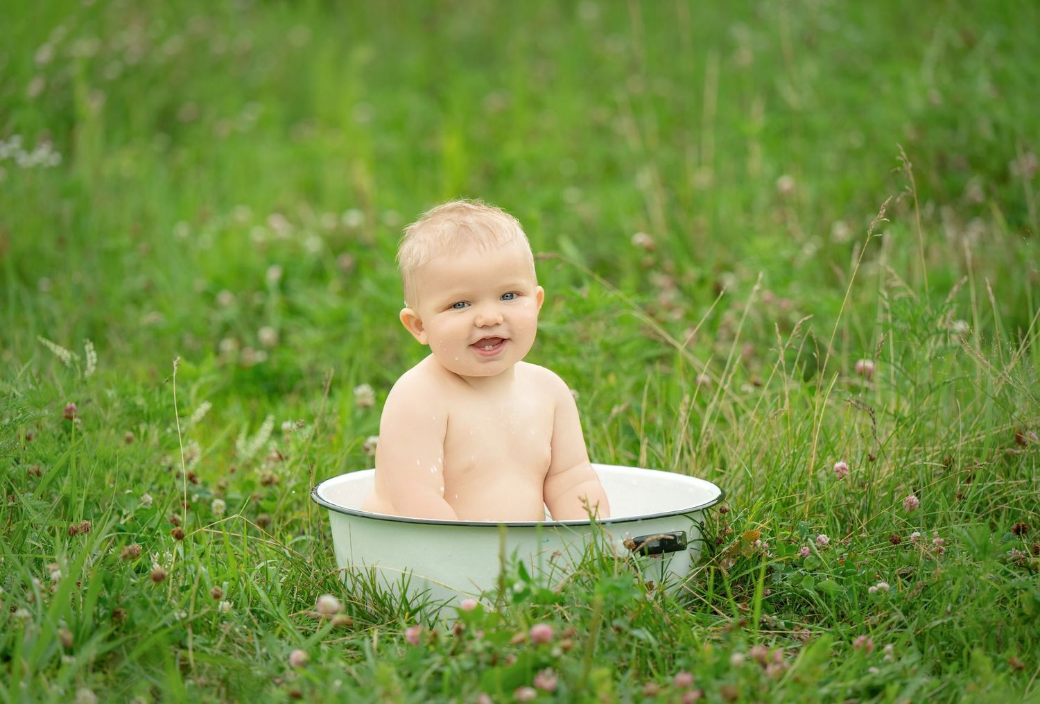 A happy baby sits in a vintage enamel wash basin surrounded by lush green grass on a sunny day. Family Portraits & Milestone Moments in Fore