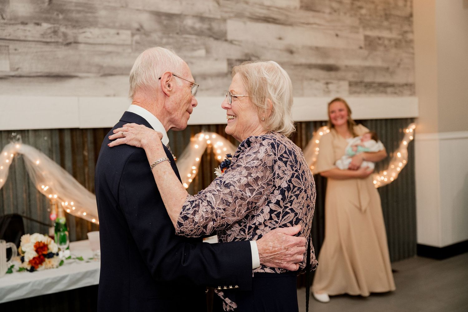 An elderly couple shares a sweet dance moment together at a wedding reception with string lights in the background.