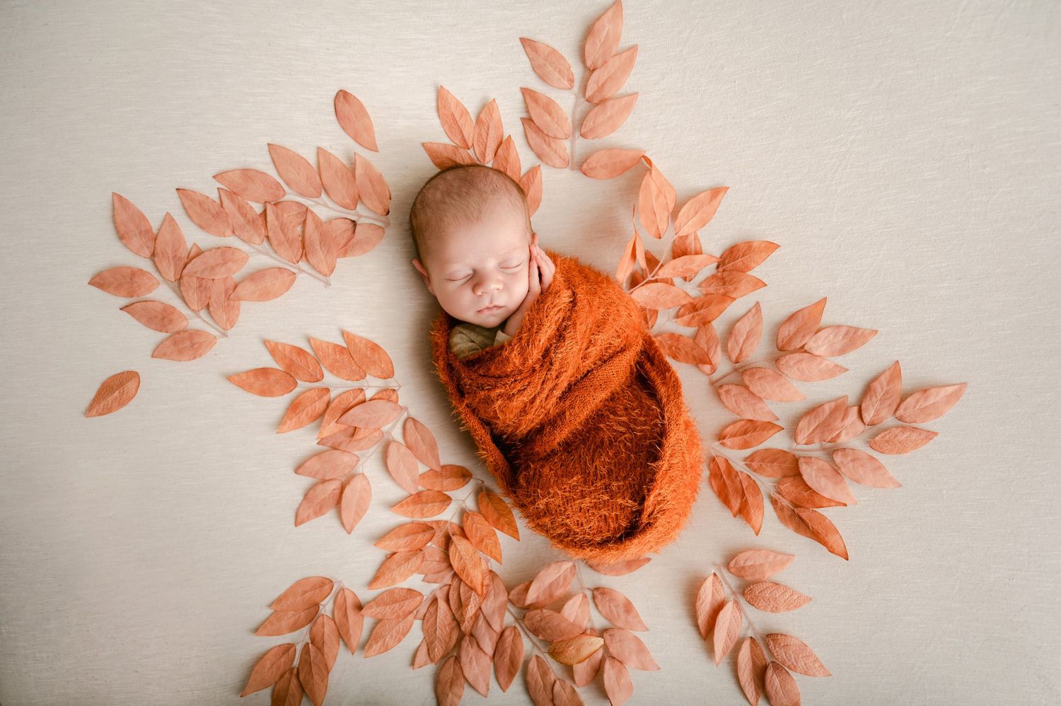 A sleeping newborn wrapped in a rust-colored swaddle blanket surrounded by delicate peach-colored leaves on beige fabric.