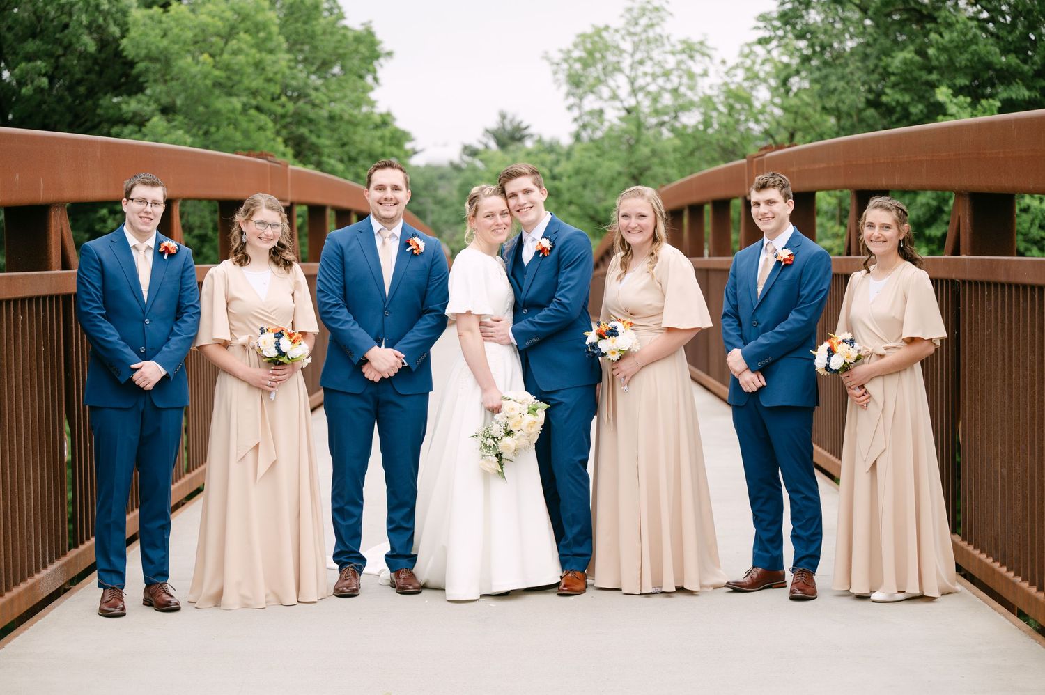 Wedding party members stand in a row on a rustic wooden bridge with alternating blue suits and blush gowns.