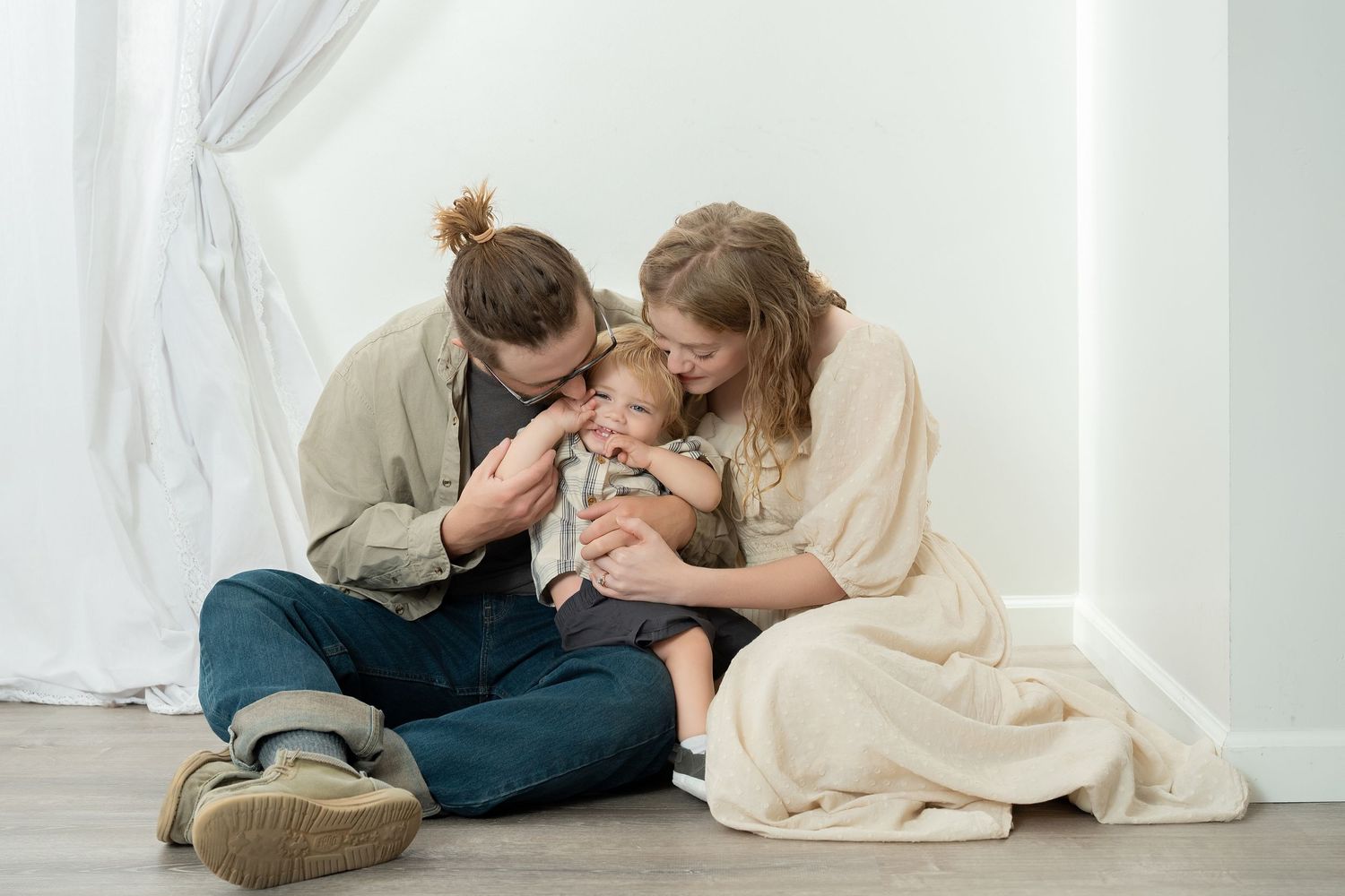 Happy family with young child sitting together on floor, parents kissing baby in bright, minimalist home setting.. Minnesota milestone photo