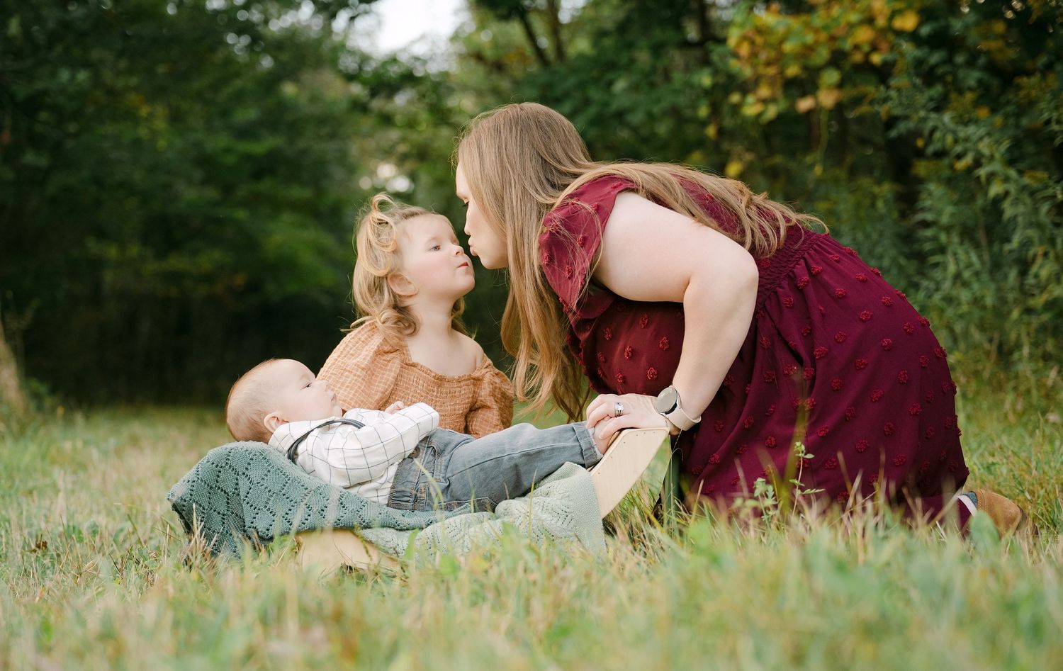 A mother shares a tender moment with her toddler and baby outdoors in a lush green field.. Minnesota milestone photographer
