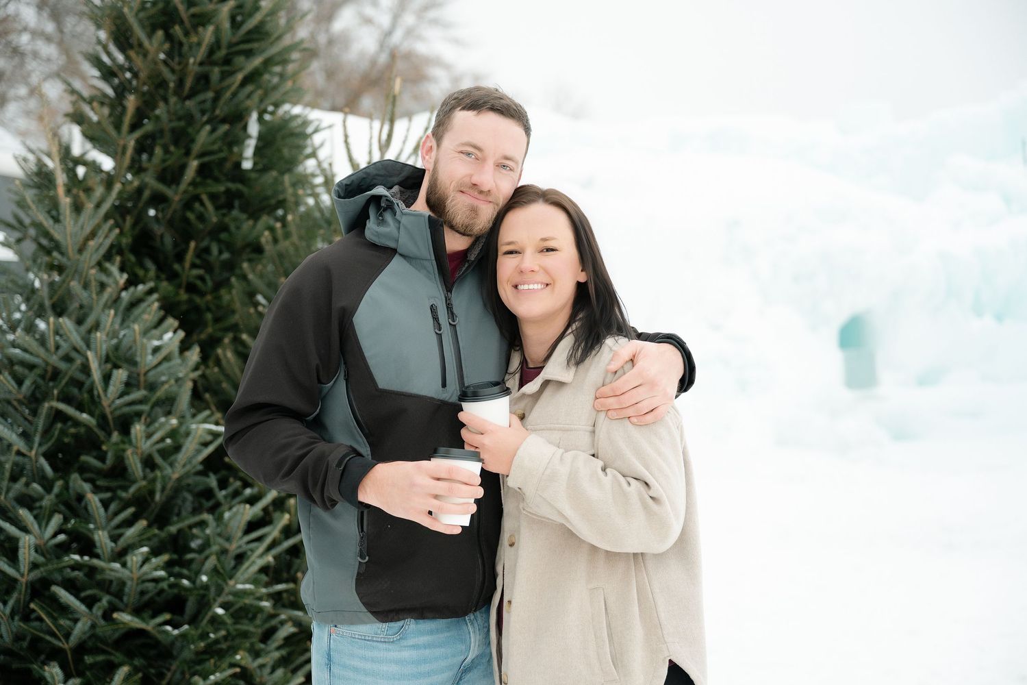 Couple smiling and holding coffee cups outdoors in winter snow near Christmas trees. Engagement Minneapolis MN