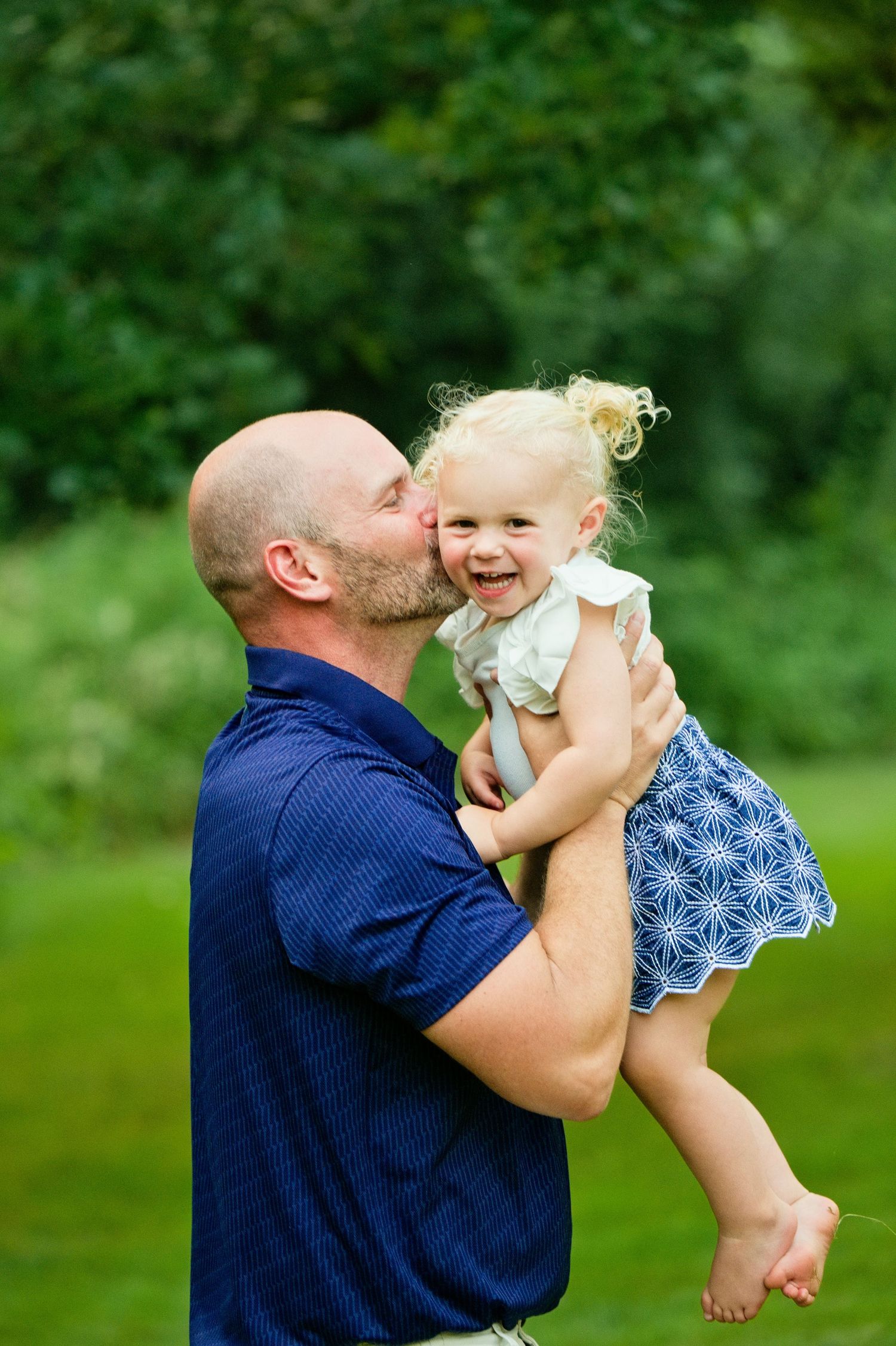 Bald man in blue polo shirt kissing cheek of laughing blonde toddler girl in blue dress outdoors.