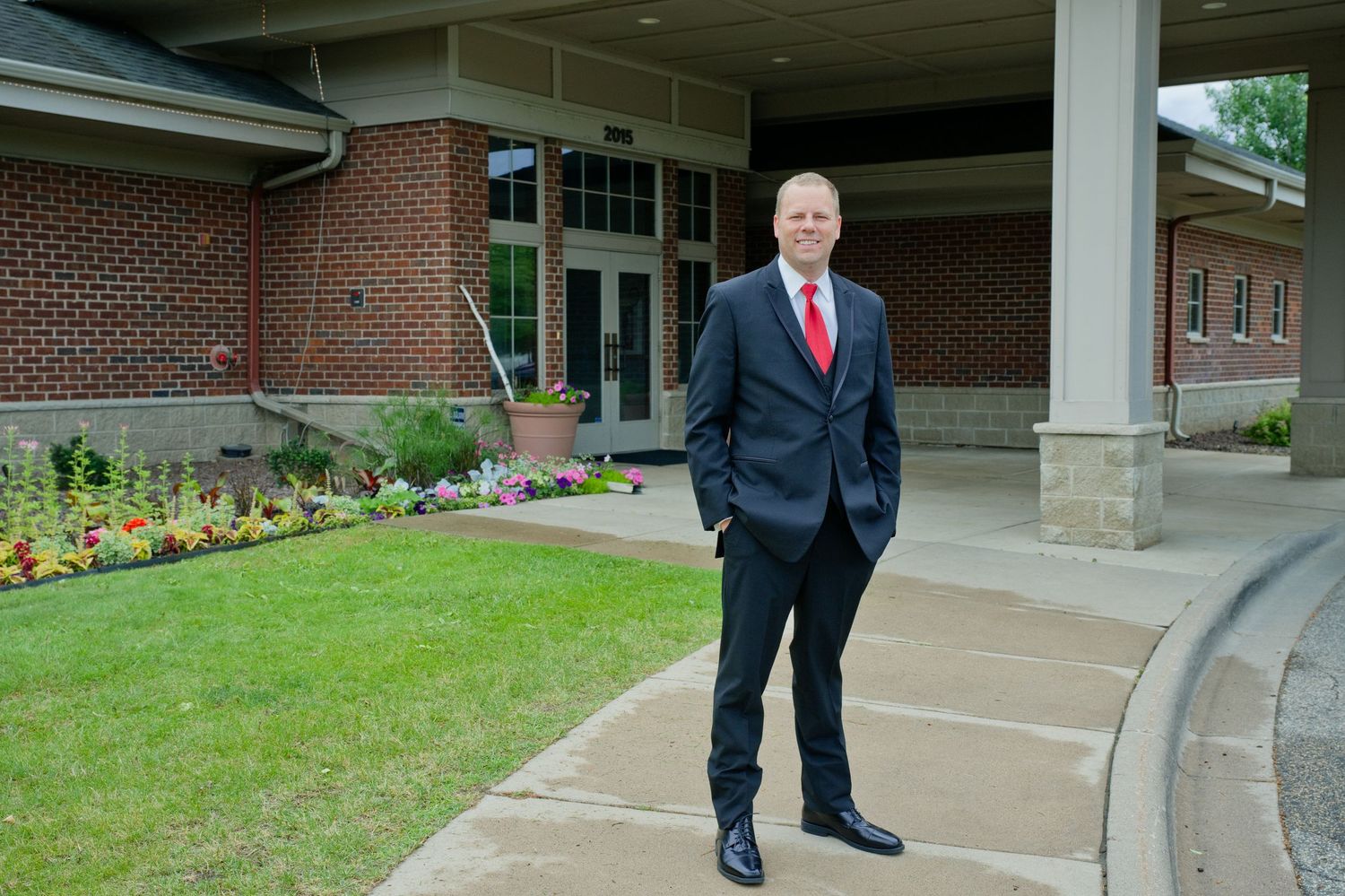 Well-dressed man in dark suit with red tie stands outside brick building with gardens.