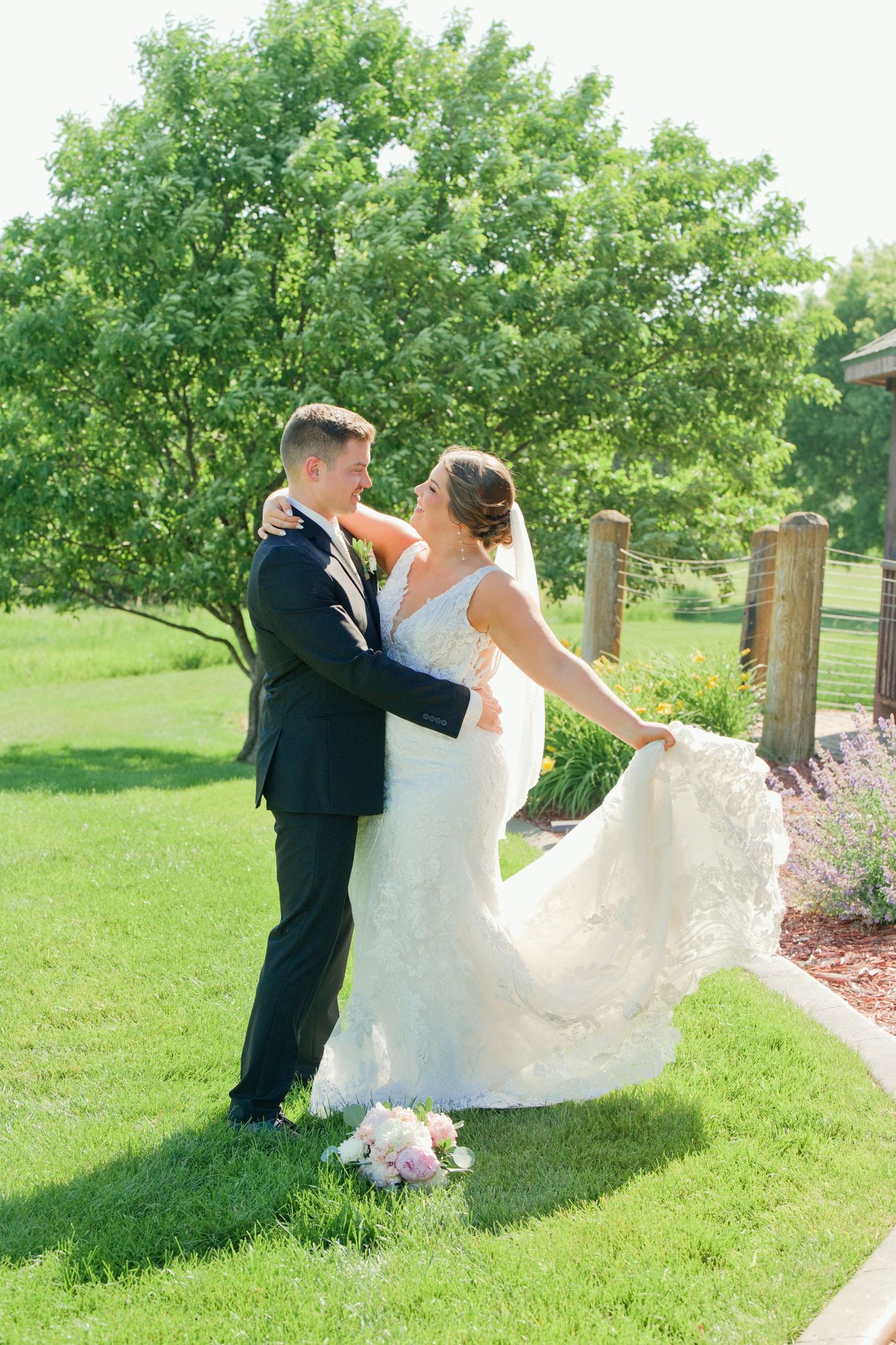 Bride in lace gown twirls with groom in navy suit on green lawn, sharing a joyful moment on their wedding day.