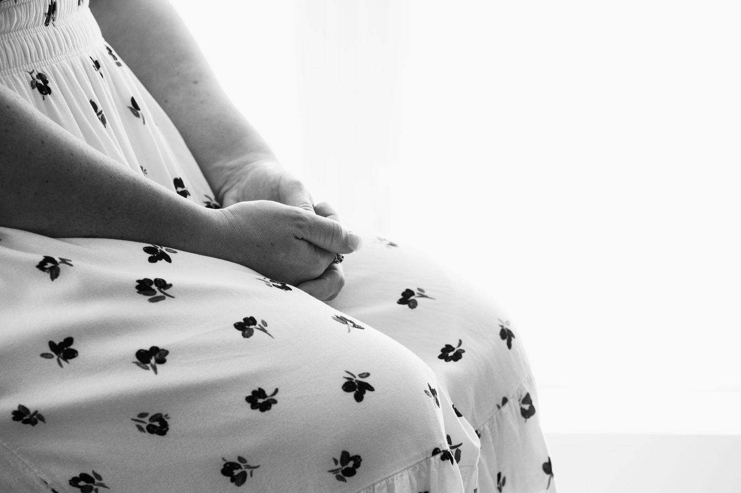 Black and white photo of pregnant woman in floral dress sitting with hands resting on baby bump.