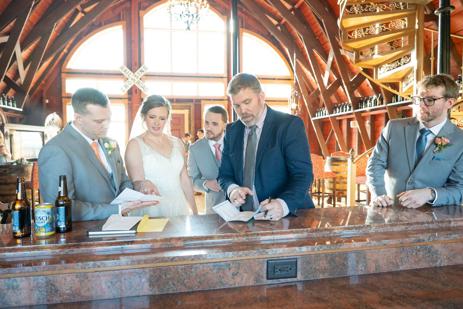 Wedding party signing marriage documents at a wooden bar in a rustic venue with arched wooden ceiling.