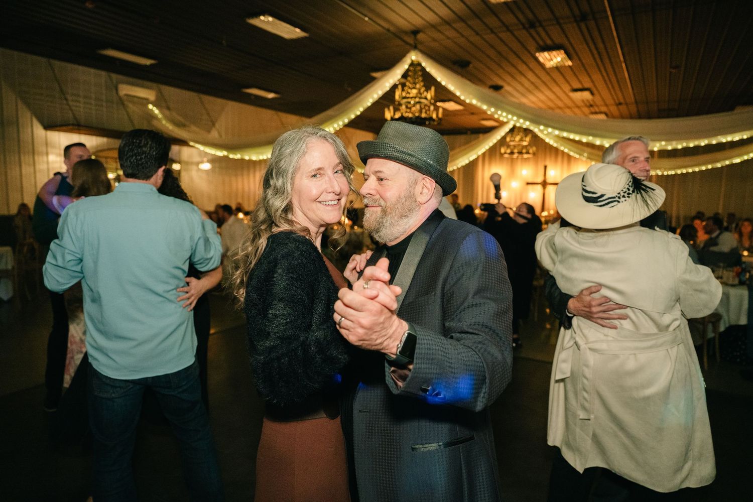 Older couple dancing joyfully at wedding reception under string lights.