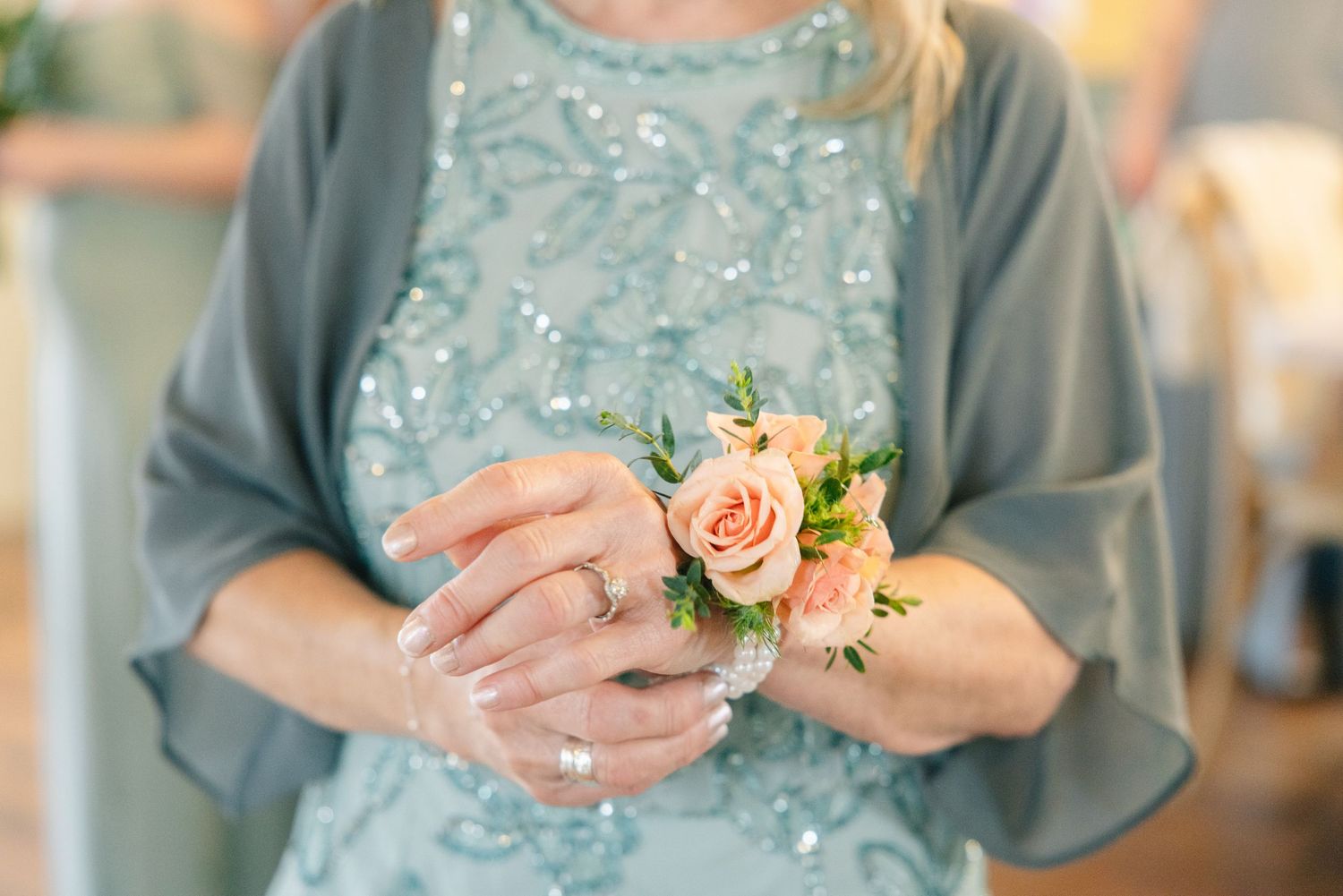 Woman in teal sequined dress holds delicate peach rose wrist corsage with manicured hands.