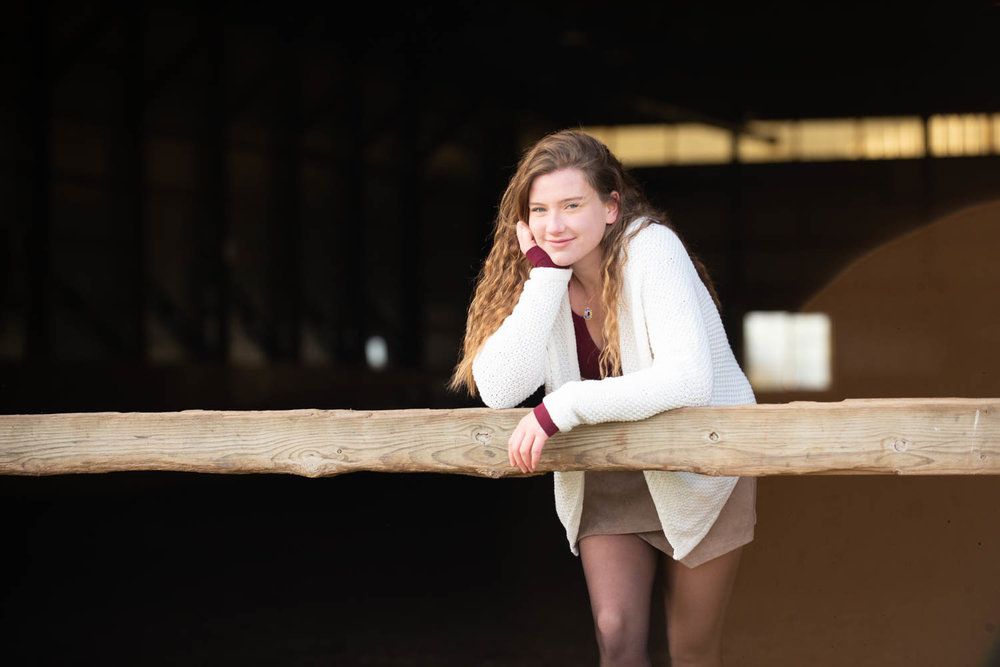 Central MN Senior Photography - Smiling young woman with curly hair leans on a rustic wooden fence rail inside a barn.