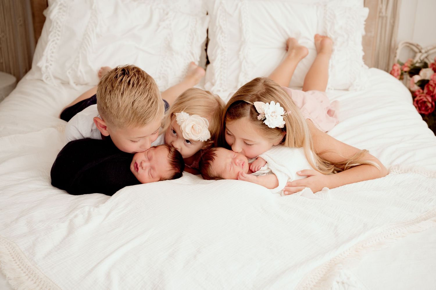 Three siblings cuddle newborn twins on a white bed, with two older kids kissing the babies in a sweet family portrait.