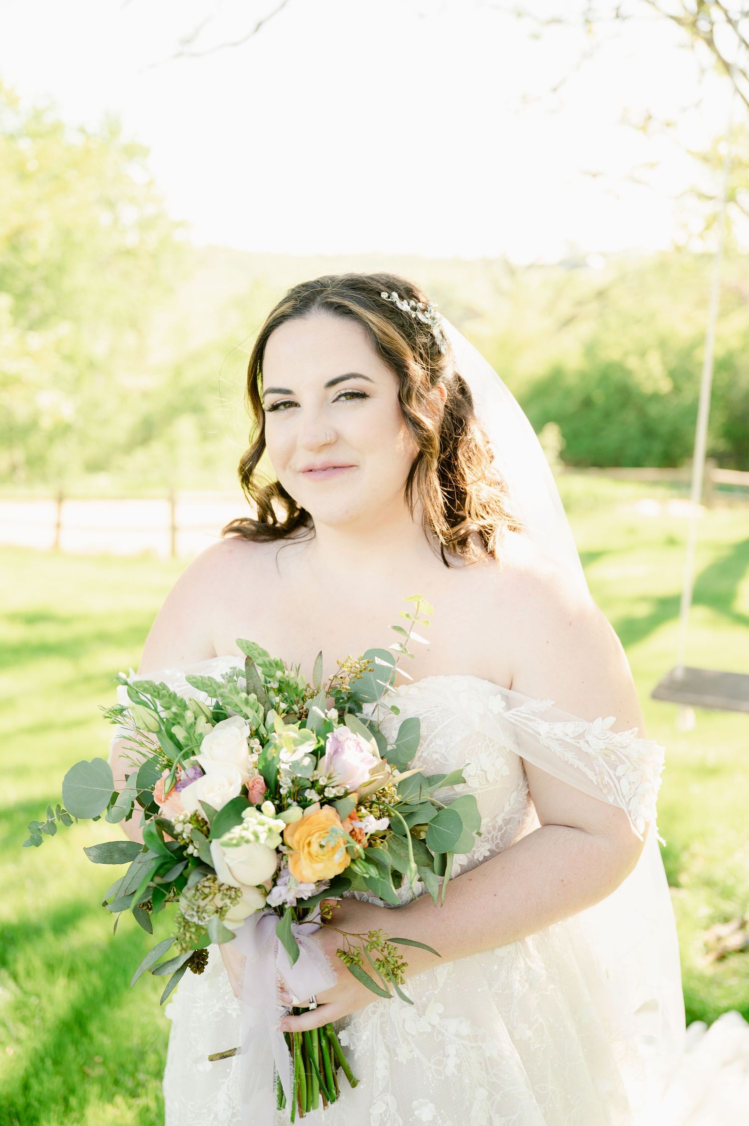 Central MN Wedding Photos - Smiling bride in lace off-shoulder gown holds pastel floral bouquet with eucalyptus in sunlit outdoor setting.