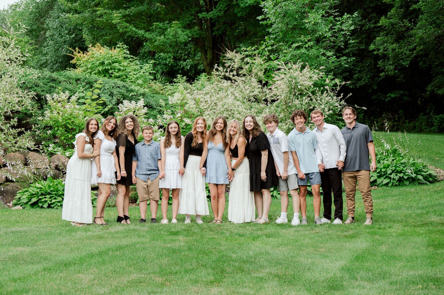 Group photo of people in light summer attire standing together on grass with lush greenery behind.