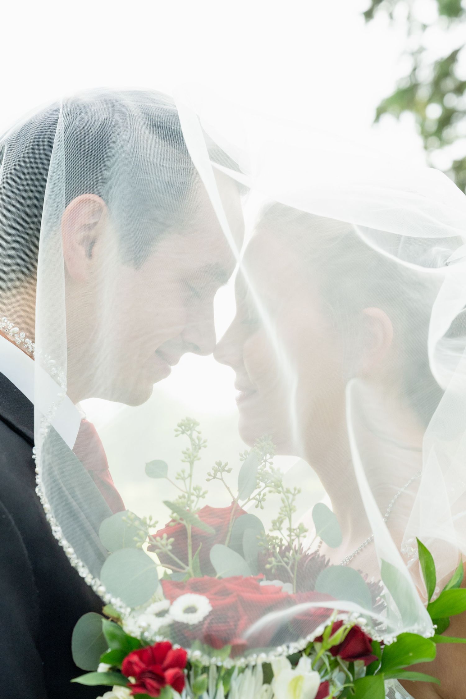 Bride and groom sharing an intimate moment under a wedding veil, holding a bouquet of red roses and greenery.
