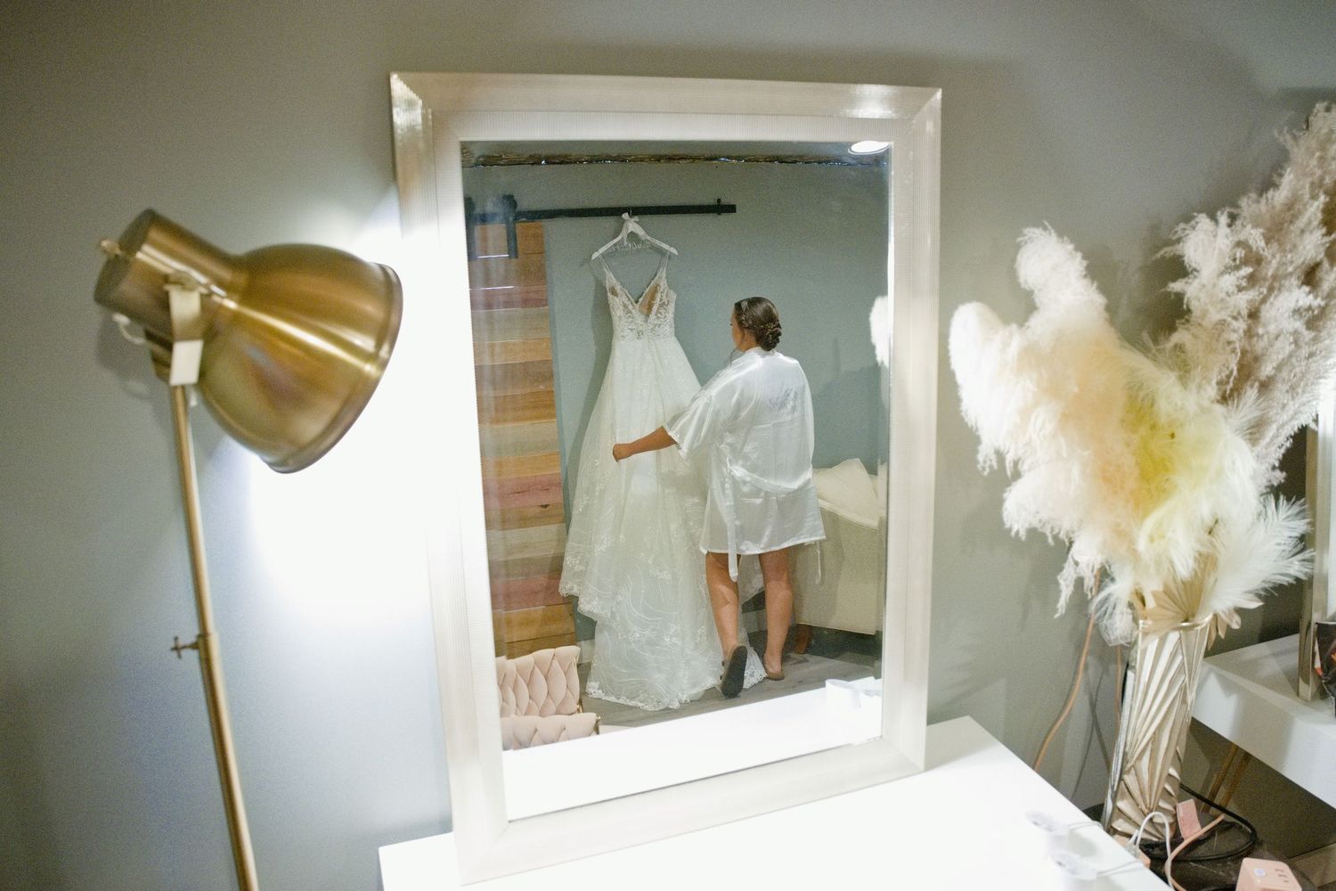 Bride reflected in a mirror admiring her wedding dress hanging in an elegantly decorated bridal suite.