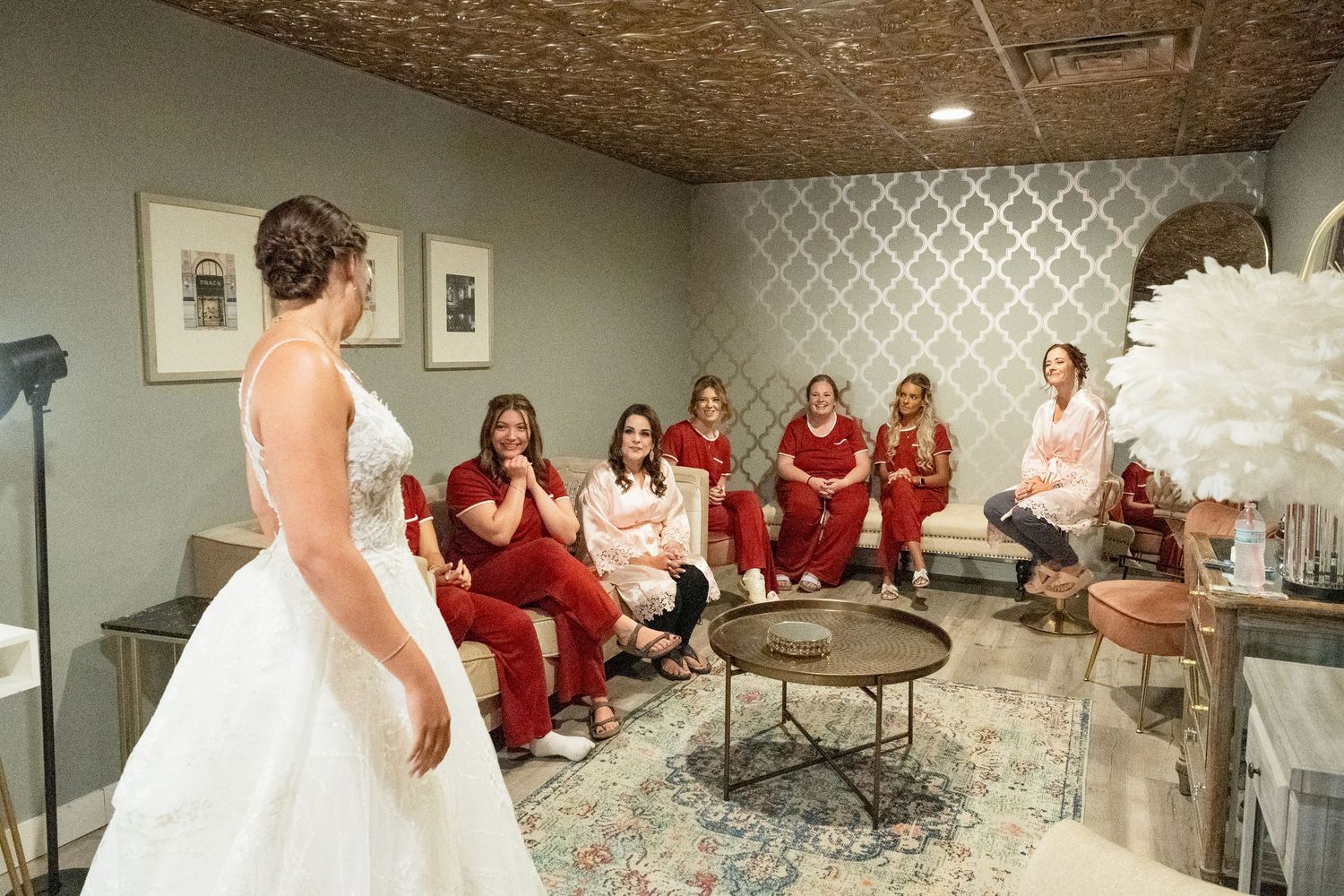 Bridesmaids in red jumpsuits react with joy as bride in white gown reveals her wedding dress in a bridal suite.