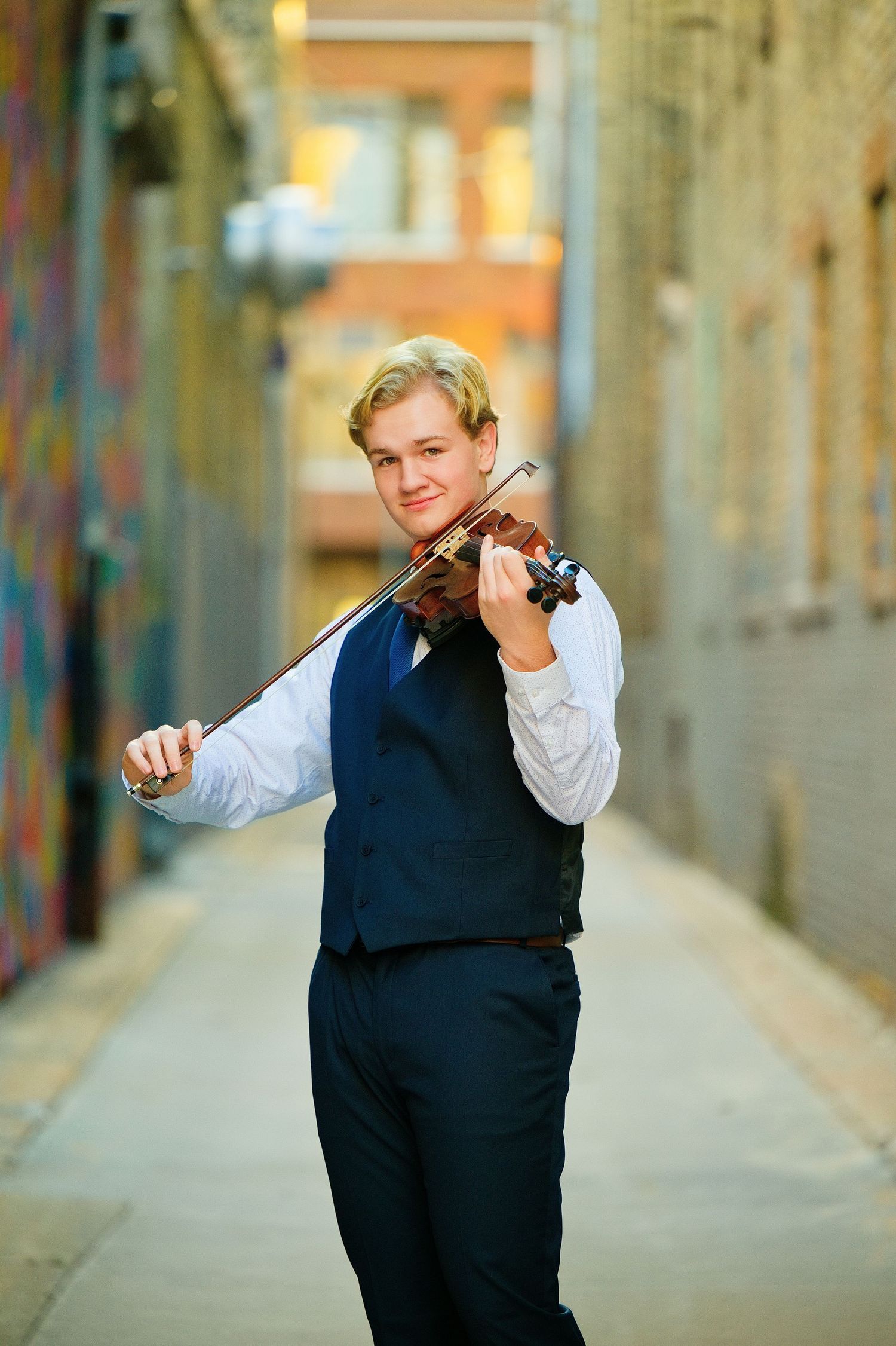 Senior Photos St Cloud MN - Young blond violinist in navy vest and white shirt plays violin in urban brick alley with colorful graffiti.