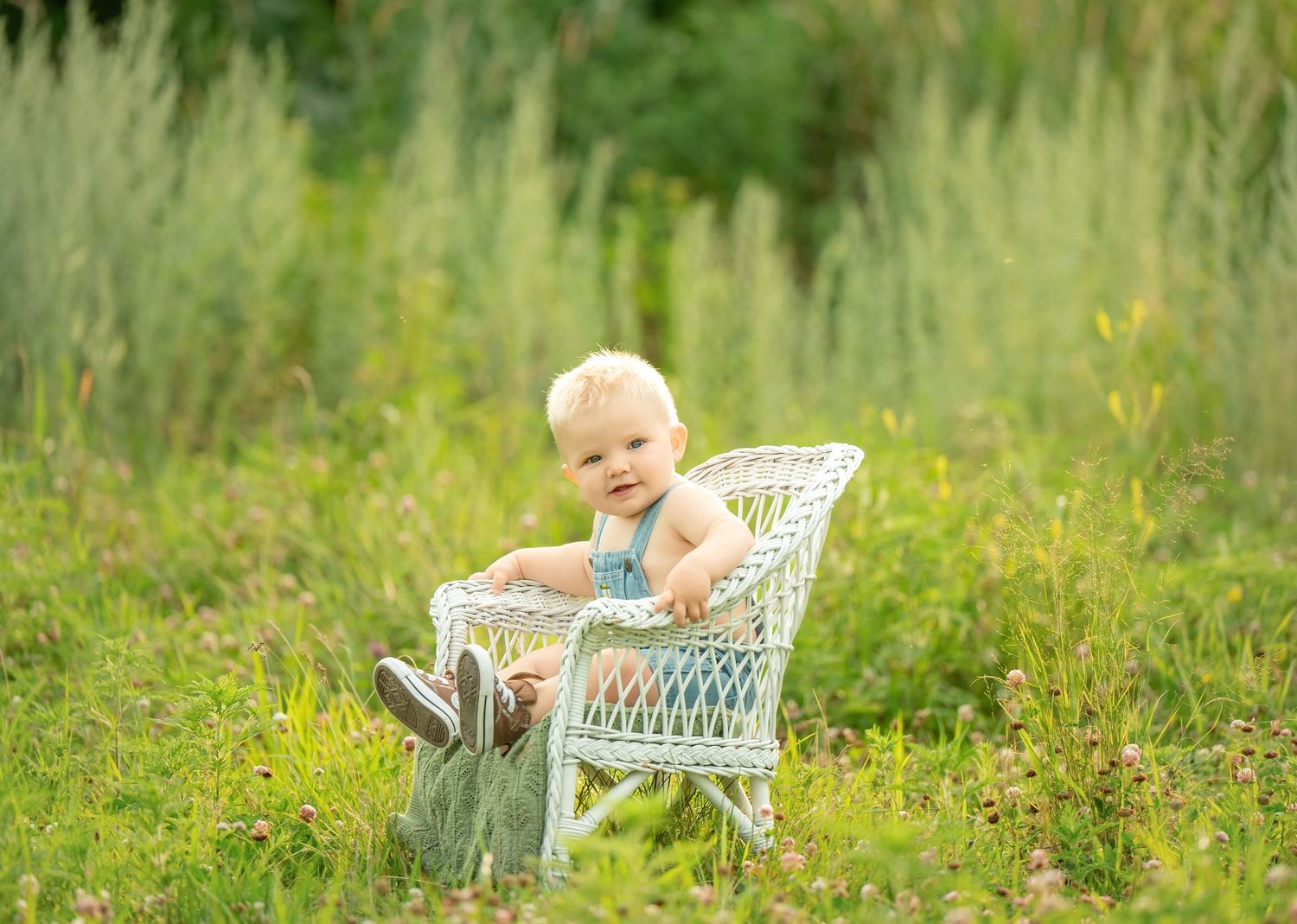 A toddler sits in a white wicker chair surrounded by tall grass and wildflowers on a sunny summer day. Family Portraits & Milestone Moments