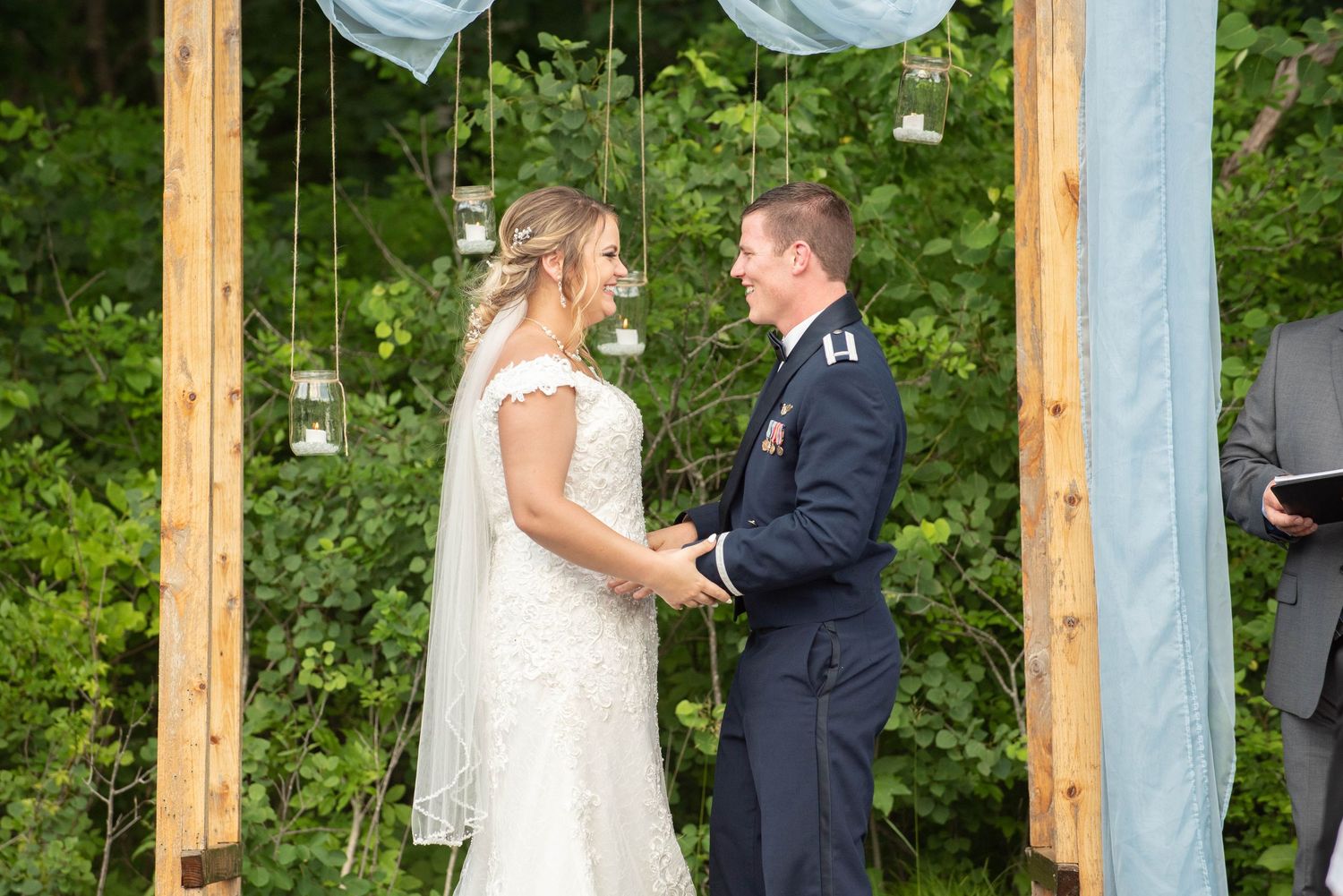 Couple exchanges vows beneath rustic wooden wedding arch with blue draping and floral decor. Zimmerman MN Weddings
