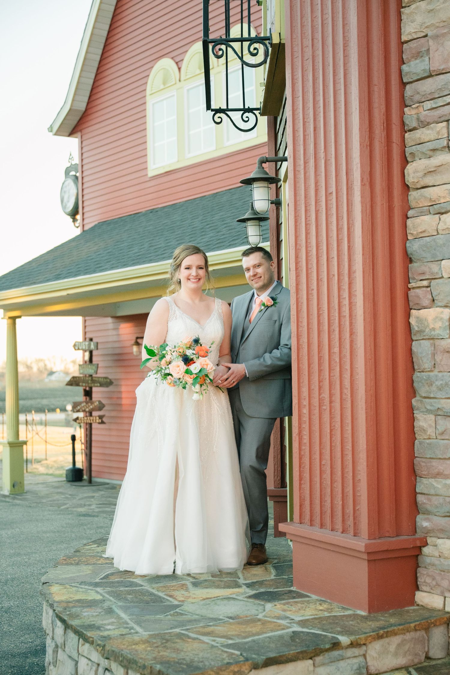 Smiling bride and groom pose outside red barn wedding venue with bride holding peach and orange bouquet.