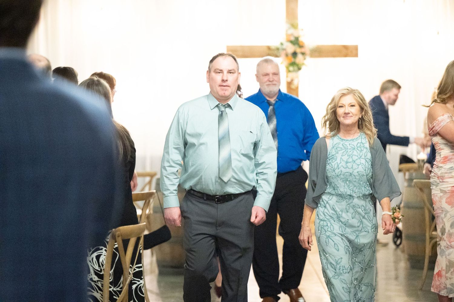 Couple in teal formalwear walk down wedding aisle past seated guests toward wooden cross altar.