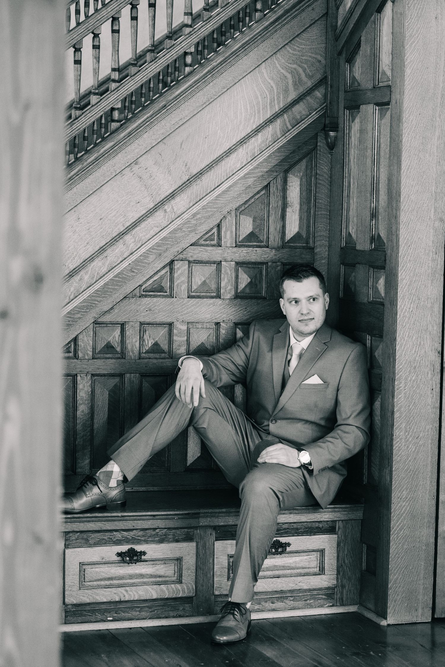 Black and white portrait of a groom in a suit sitting relaxed beneath an ornate wooden staircase.