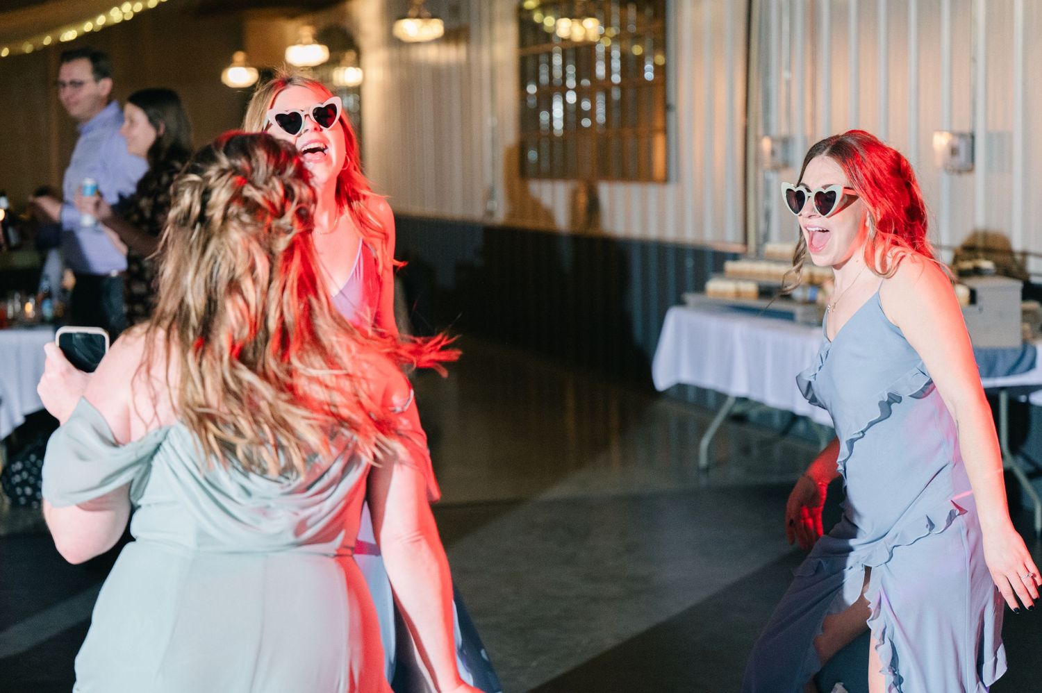 Wedding guests wearing heart-shaped sunglasses dance energetically under red lights at the reception.
