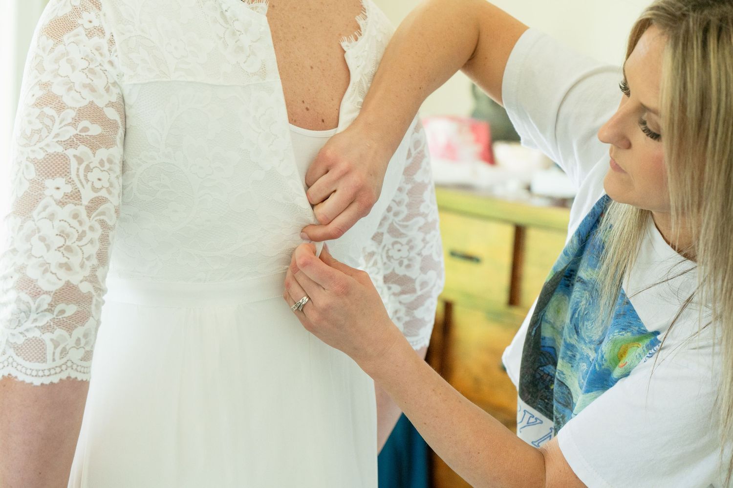 A woman in a white lace wedding dress gets helped with buttons by a blonde woman in a blue floral top.