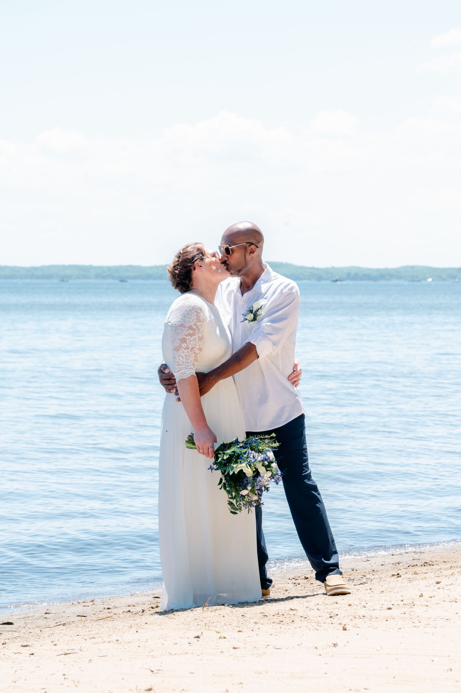 Bride and groom sharing a kiss on a sandy beach with calm blue water in the background on their wedding day.