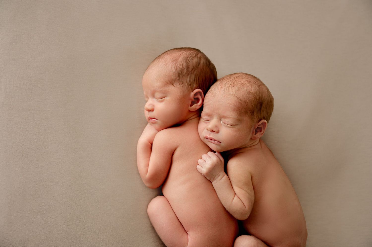Two sleeping newborn twins cuddled together on a soft beige background in a peaceful studio portrait.