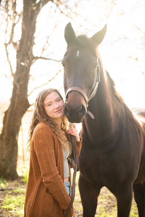 Central MN Senior Photography - Young woman with braided hair in brown cardigan stands beside a dark bay horse outdoors in golden sunlight.