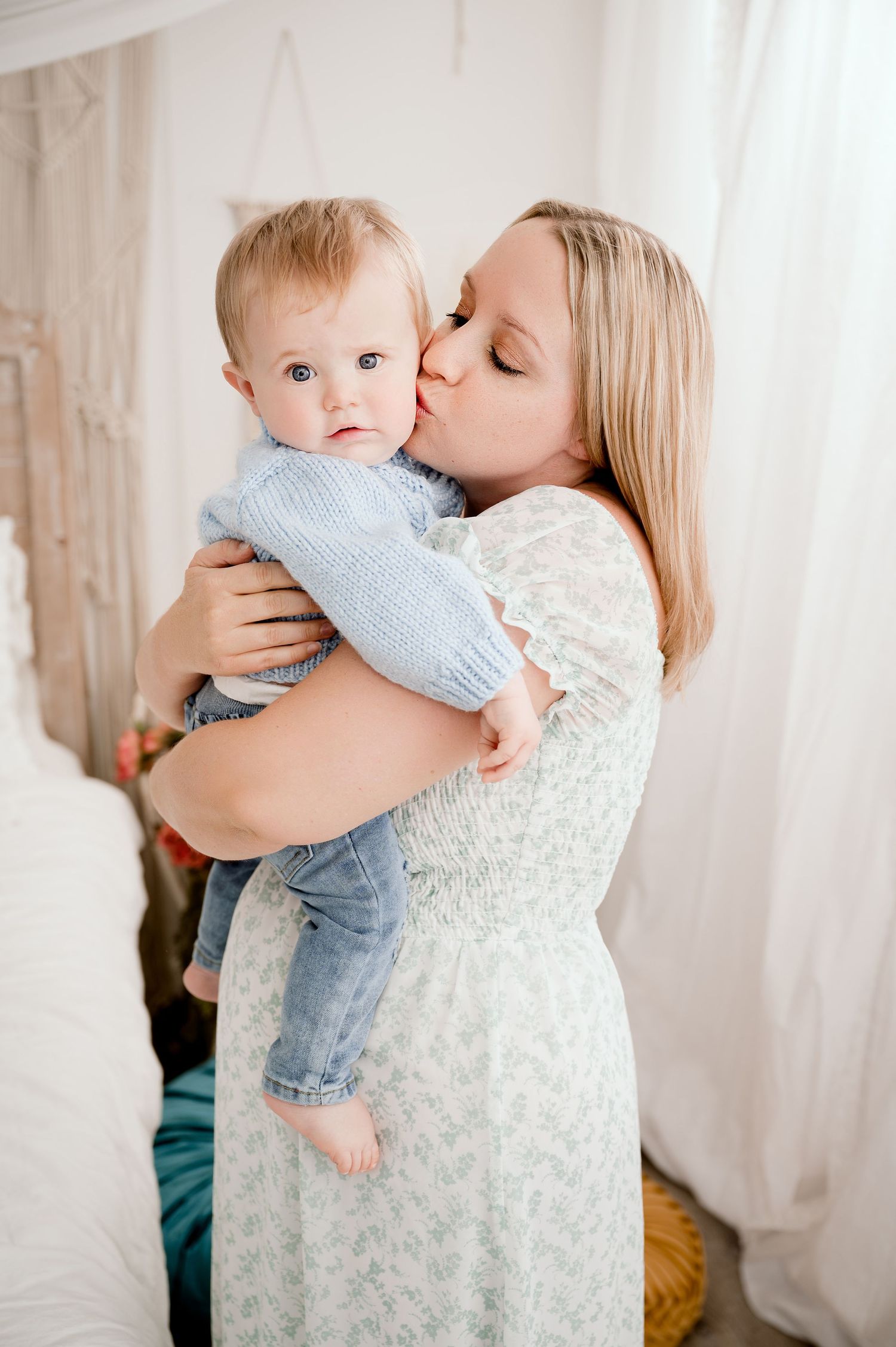 Central MN Milestone Photography - Mother in floral dress kissing her baby on the cheek in a bright white boho bedroom.