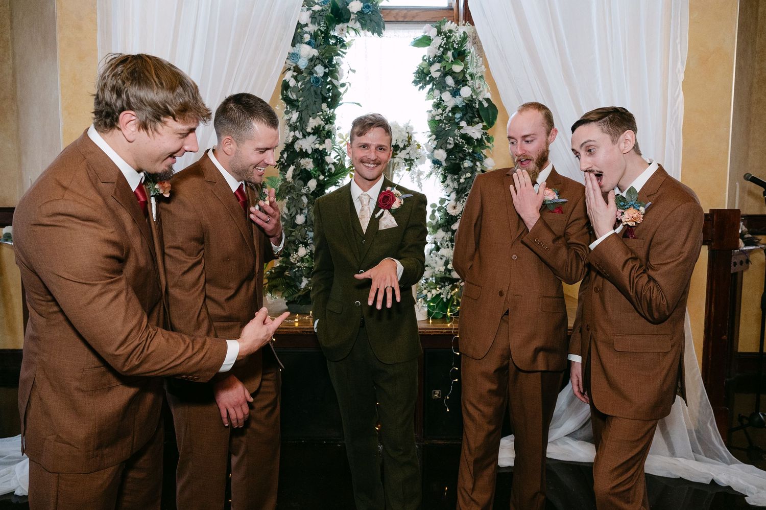 Central MN Wedding Photos | Groom in olive suit laughing with groomsmen in brown suits near decorated wedding ceremony arch.