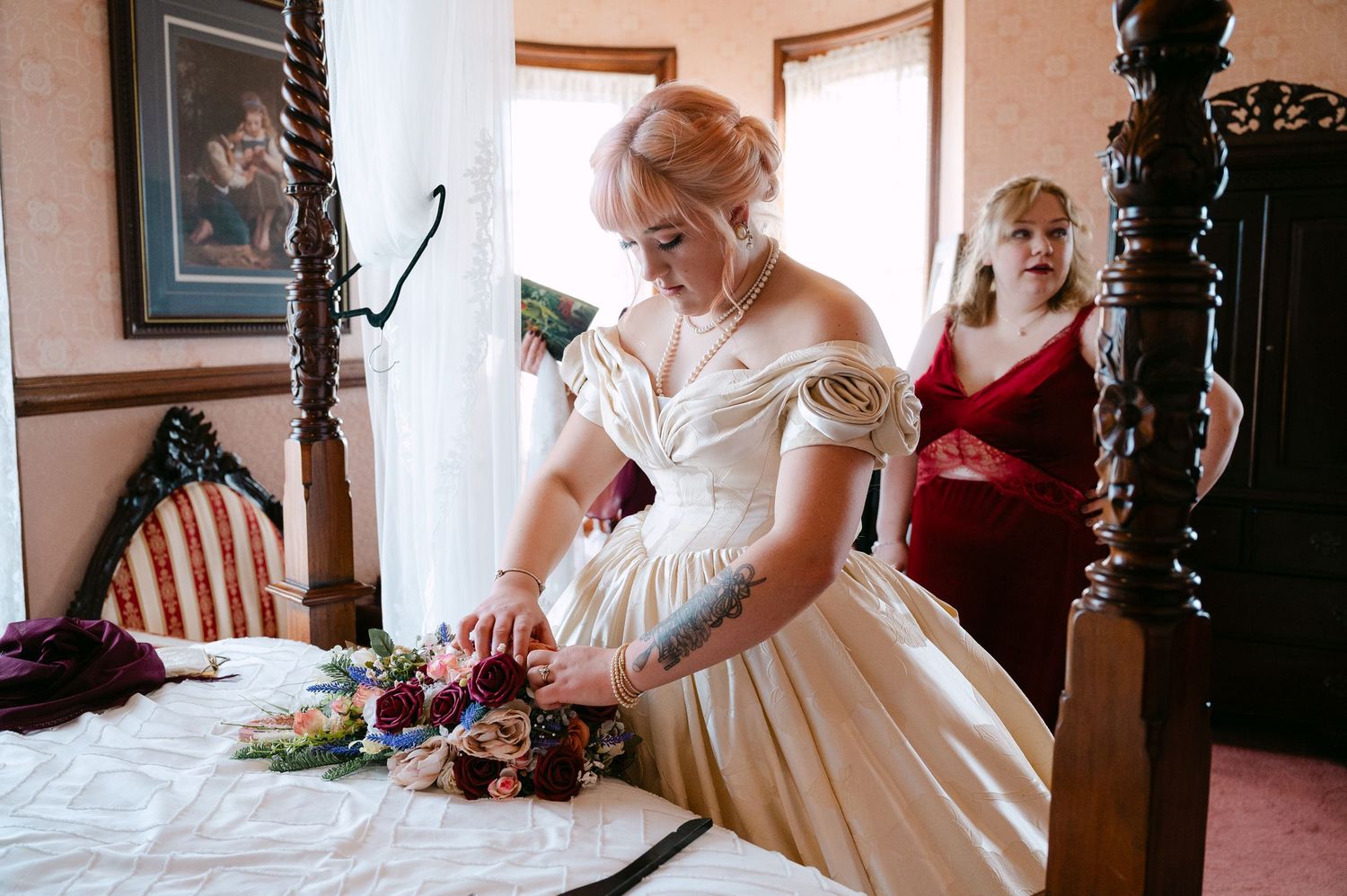 Central MN Wedding Photos | Bride in off-shoulder ivory ball gown arranging bouquet on bed with bridesmaid in red dress nearby.