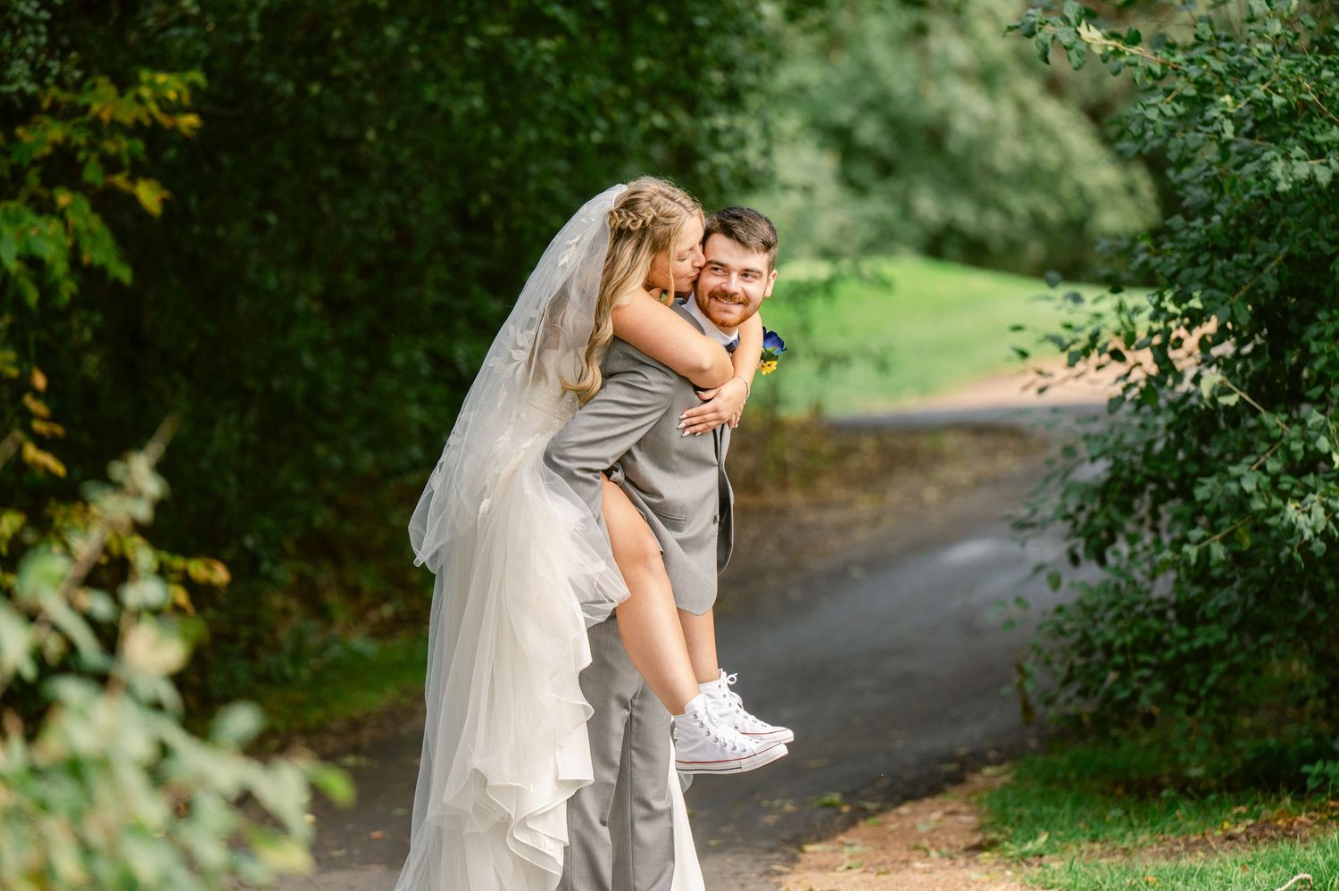Central MN Wedding Photos | Groom gives bride a playful piggyback ride along a lush green garden pathway outdoors.