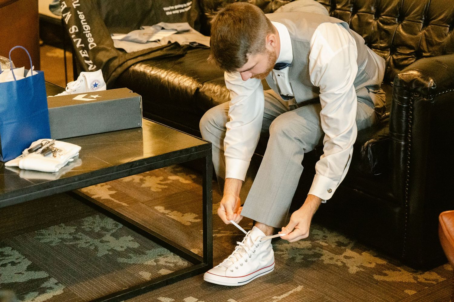 Central MN Wedding Photos | Groom in gray suit and navy bow tie laces up white Converse sneakers before wedding ceremony.