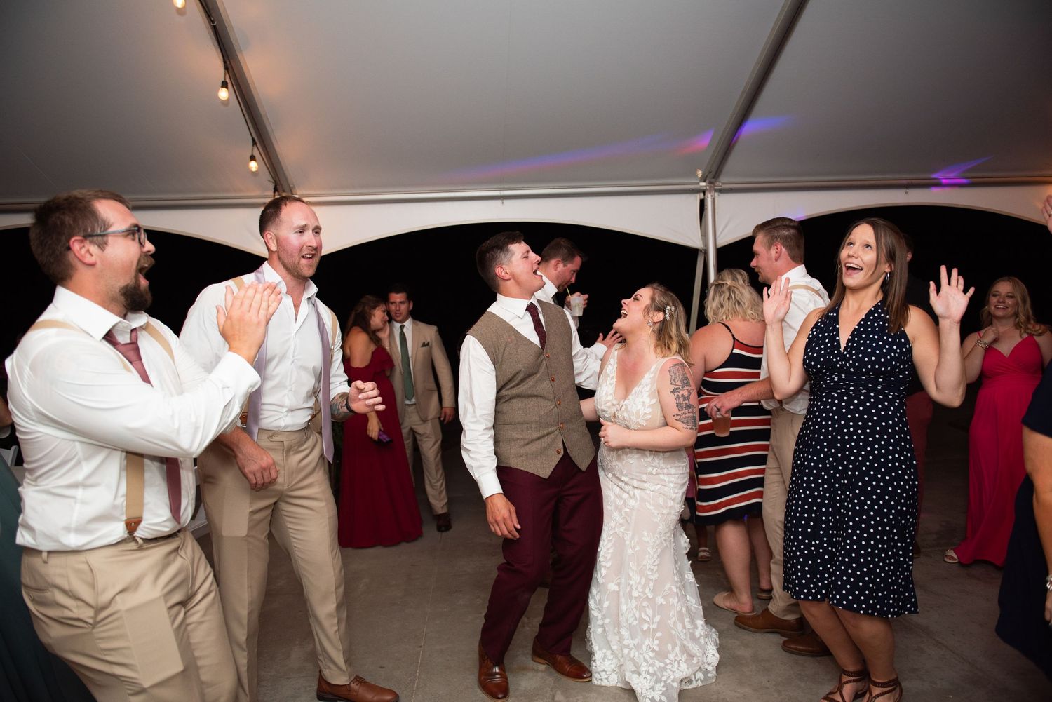 Reception guests dancing together under a white tent canopy with colored lights creating a festive atmosphere.