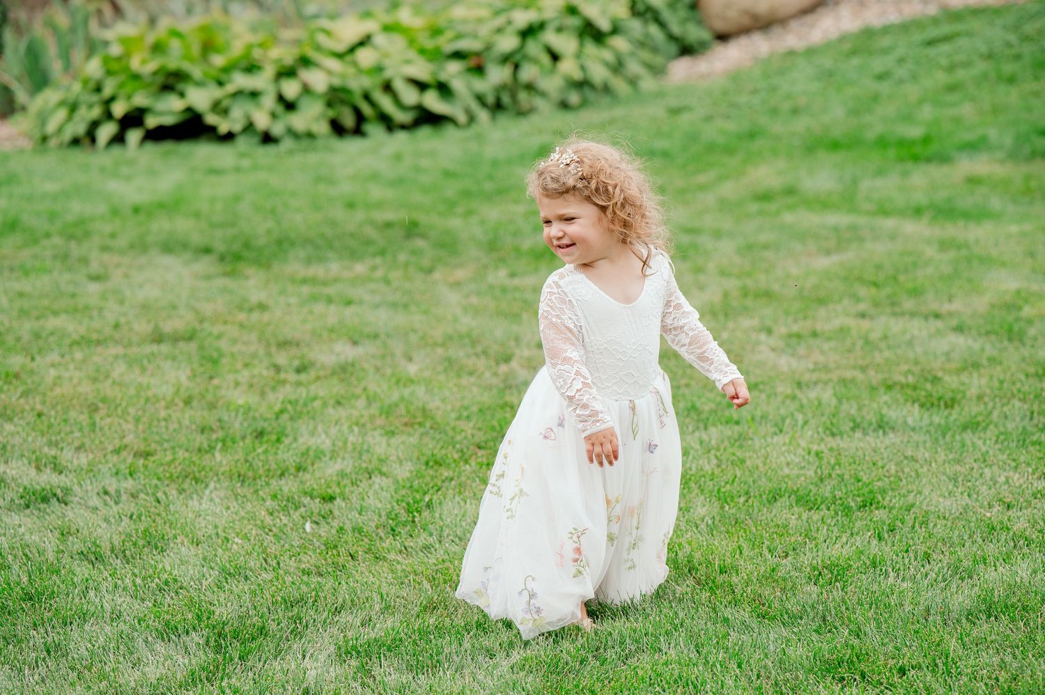 Young flower girl in white lace dress plays on green lawn.