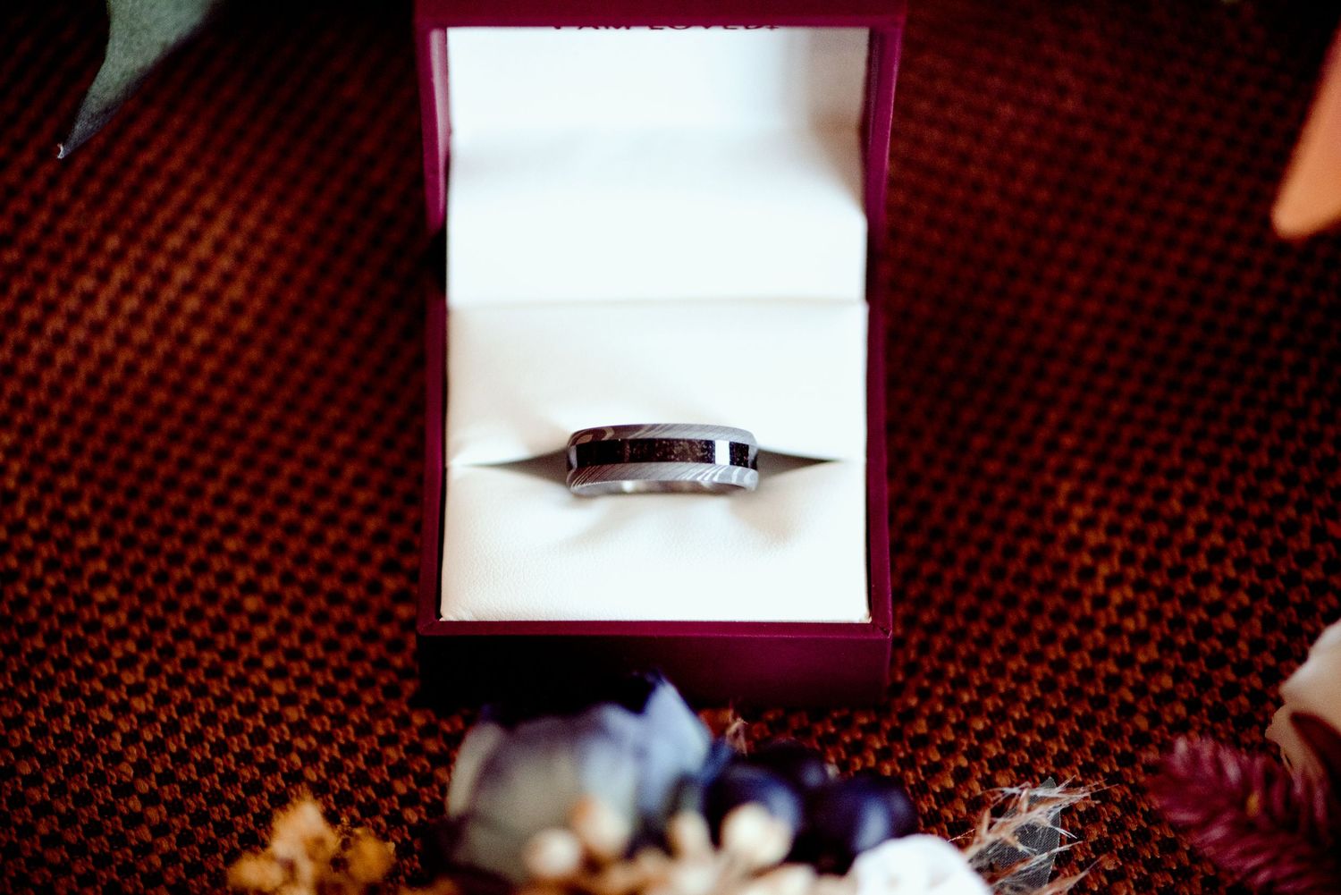 A single diamond engagement ring sits in a pink velvet ring box on a deep red textured surface.