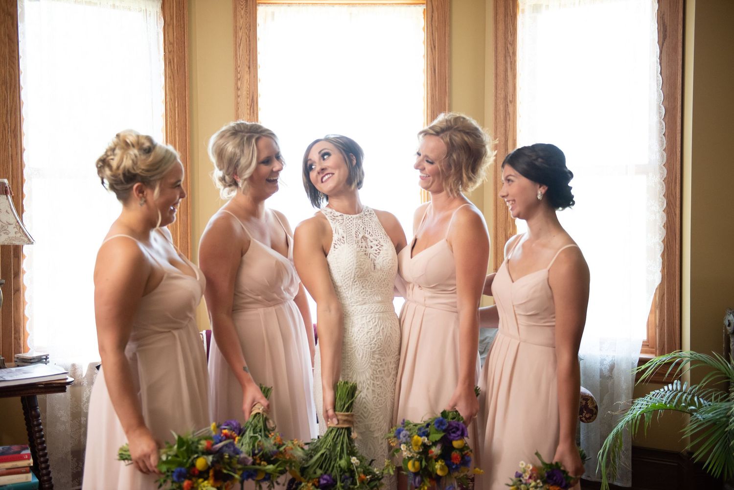 Bridesmaids in blush pink dresses stand together by window, sharing joyful moments before wedding ceremony.
