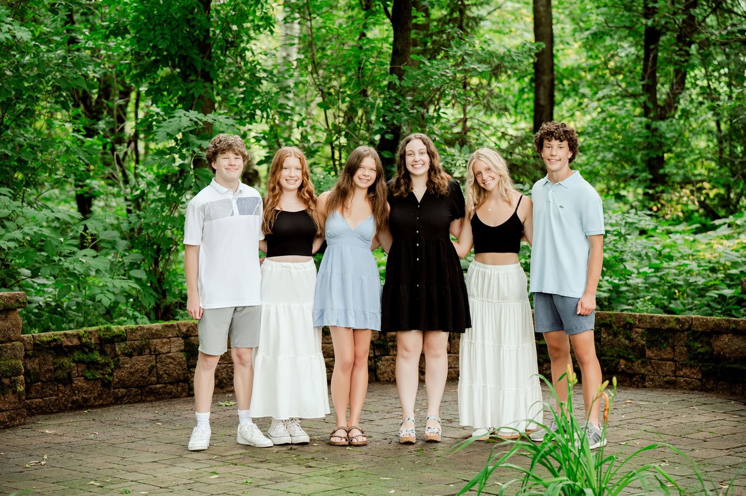 Six people in coordinated black and white outfits pose on a stone patio in a forest setting.