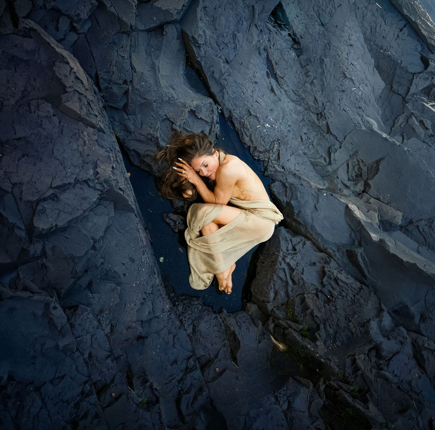 A woman in a white dress lies curled among dark blue rocky cliffs, photographed from above.
